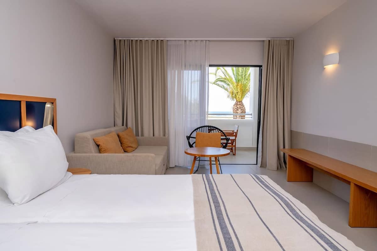 Habitación de hotel con cama, sofá, una pequeña mesa con sillas, un banco y vista a una palmera a través de la ventana.