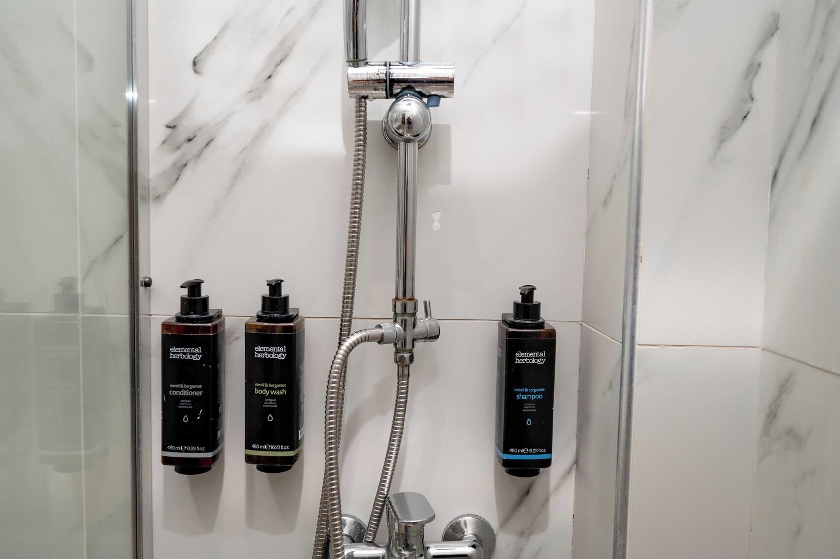 A bathroom with a shower, a handheld showerhead, and three bottles of Elemental Herbology body wash, conditioner, and shampoo.