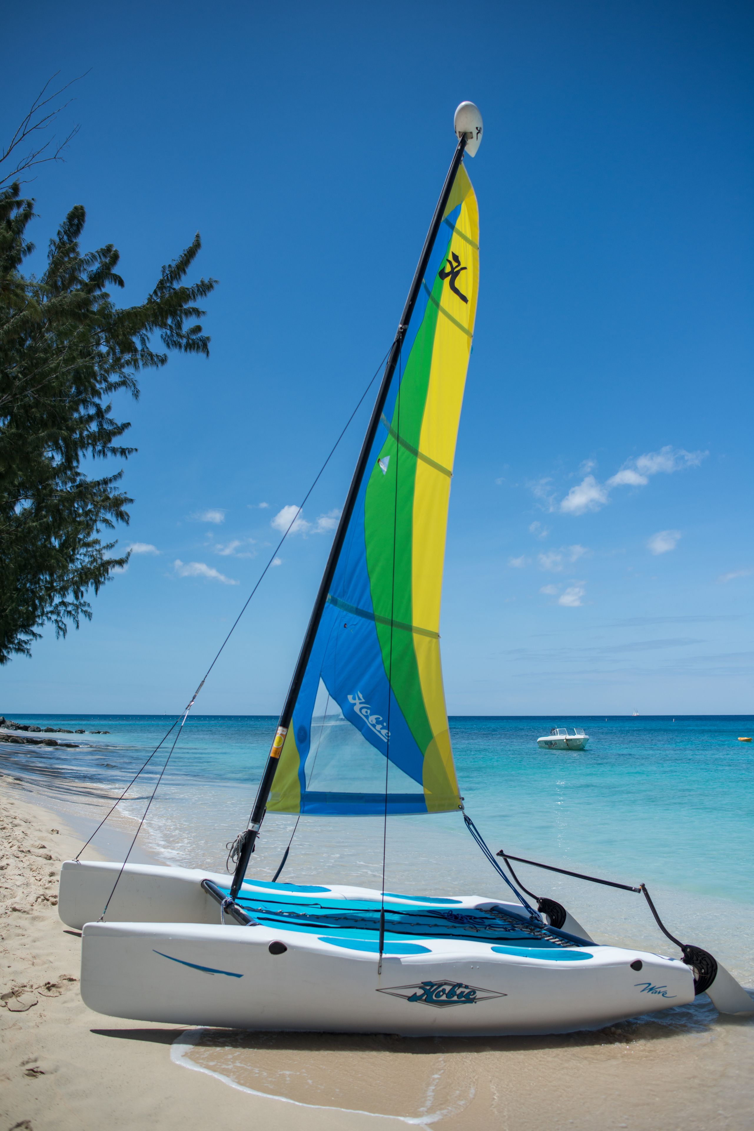 A sailboat with a blue and yellow sail on a sandy beach with clear blue water and a small boat in the distance.