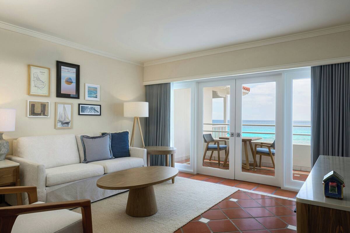 A hotel room with a balcony, a sofa, a coffee table, and a dining area with a view of the ocean.