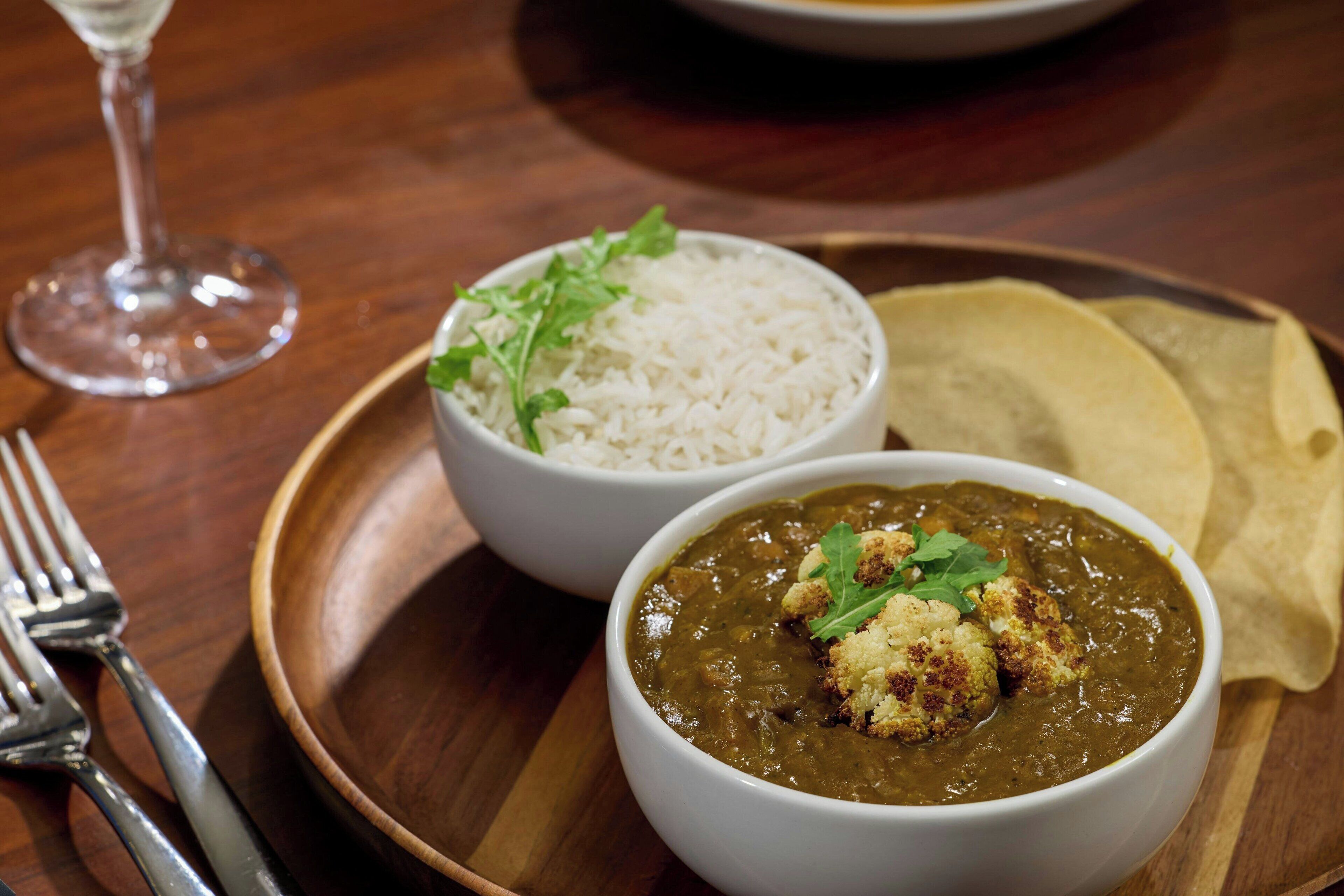 Taula de fusta amb un plat que conté un bol de curry, un bol d'arròs i un tros de pa naan.