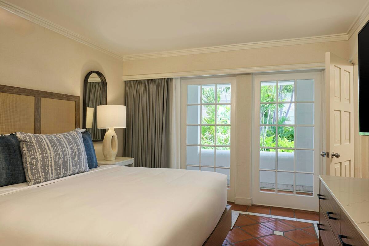 A hotel room with a large bed, a nightstand with a lamp, a mirror, and a sliding glass door leading to a balcony with greenery outside.