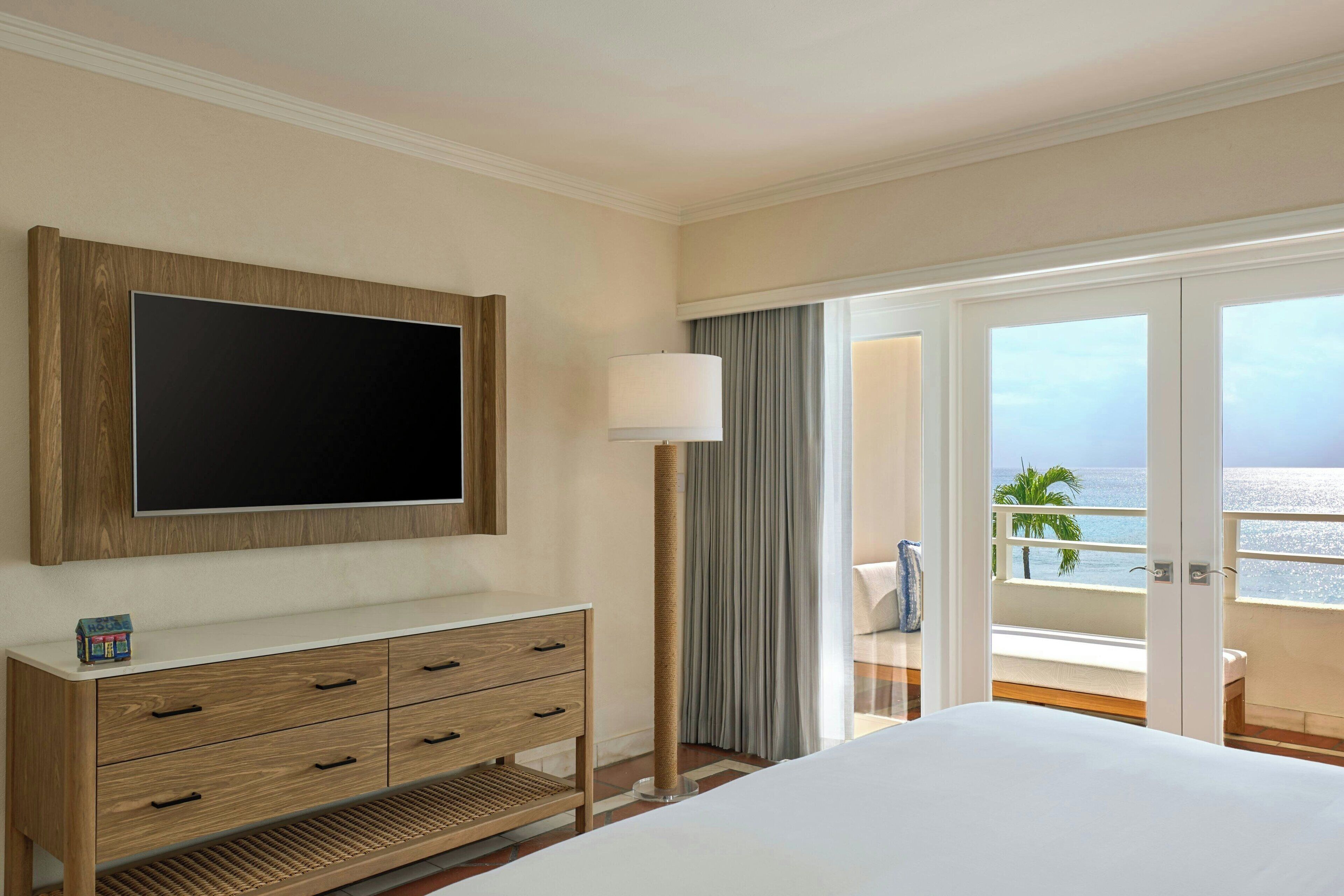 A hotel room with a large flat-screen TV mounted on the wall, a wooden dresser with drawers, a large bed, and a balcony with a view of the ocean.