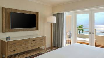 A hotel room with a large flat-screen TV mounted on the wall, a wooden dresser with drawers, a large bed, and a balcony with a view of the ocean.