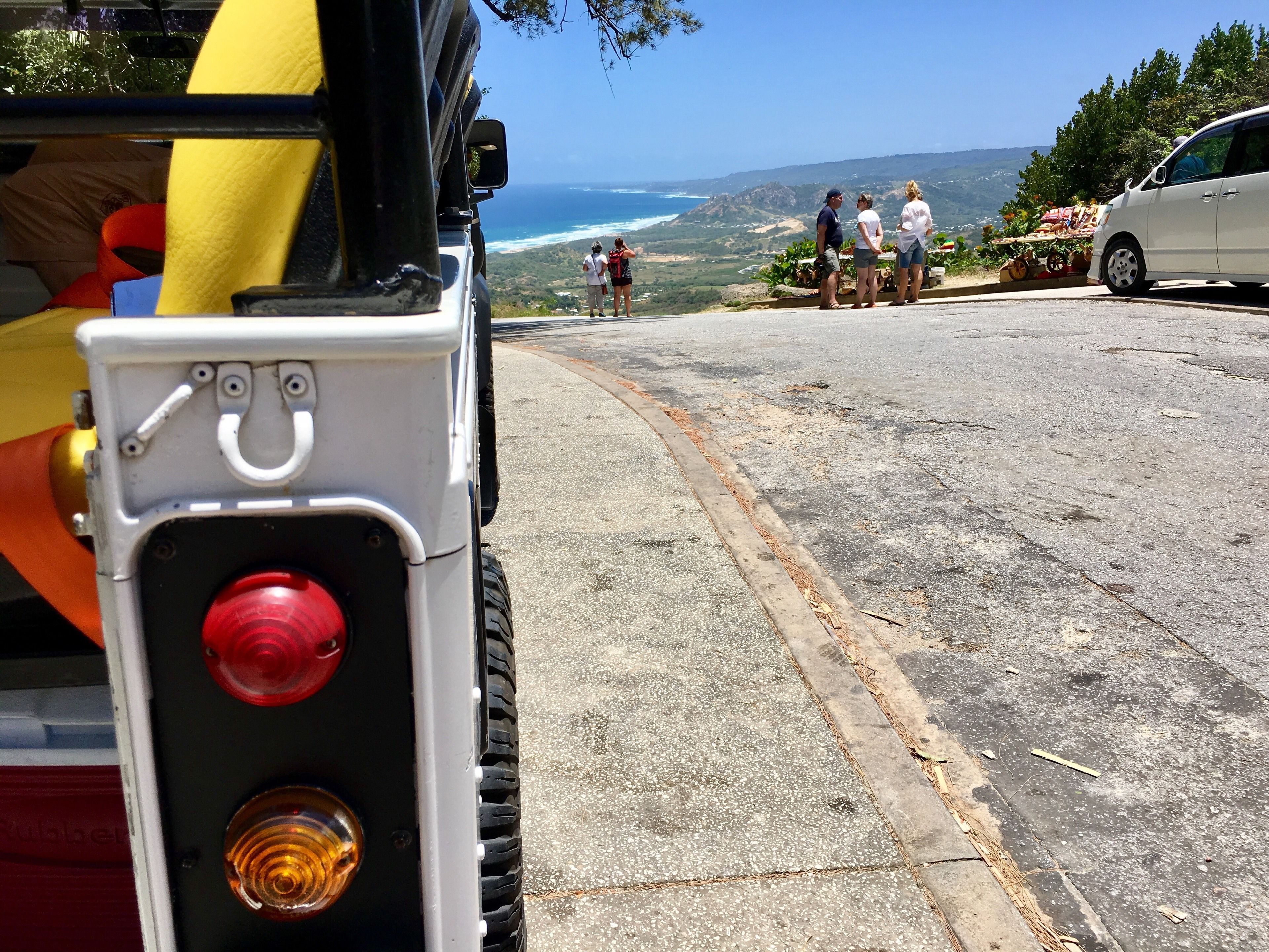 A vehicle with a red light and a yellow object, a paved road, and a group of people overlooking a coastal landscape.