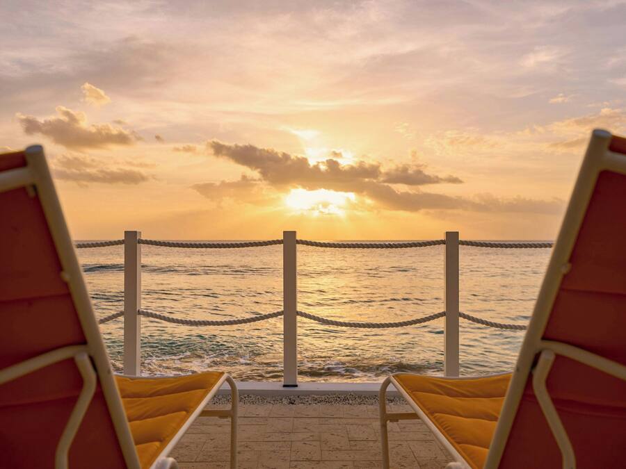 Vista de la playa con dos sillas reclinables amarillas orientadas al océano, una barrera de cuerda y un atardecer sobre el agua.