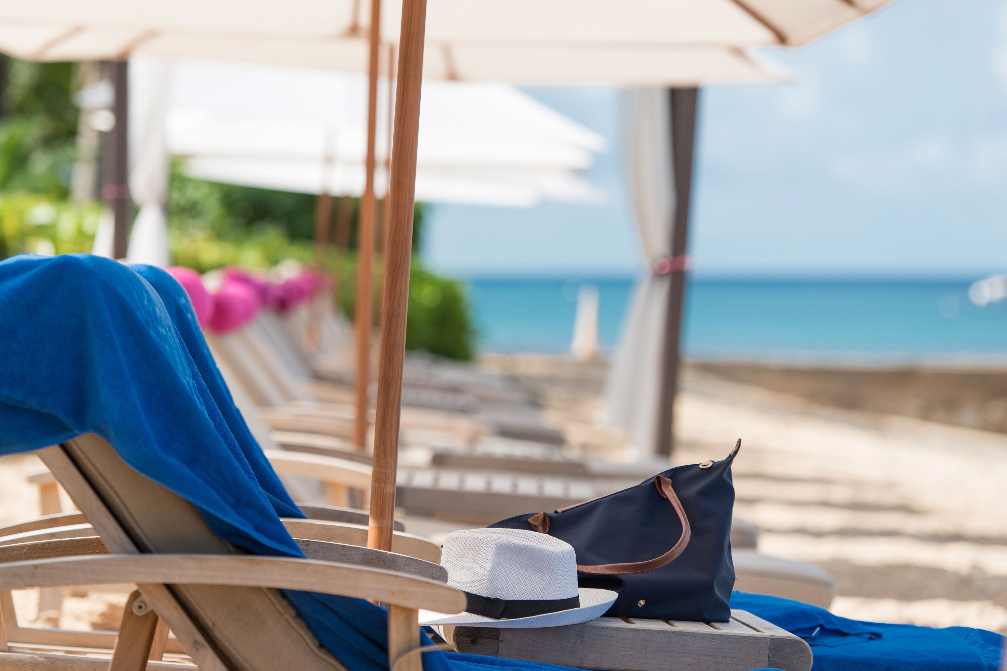 A row of wooden lounge chairs with blue towels and a white hat on one, and a navy blue bag on another, along a beachside walkway.
