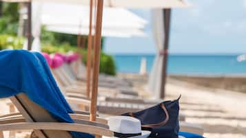 A row of wooden lounge chairs with blue towels and a white hat on one, and a navy blue bag on another, along a beachside walkway.