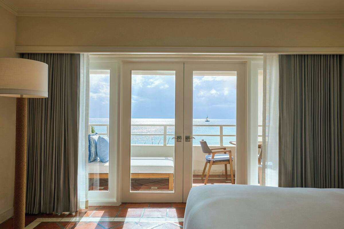 A hotel room with a bed, a chair, a view of the beach, and a balcony with a table and chairs.