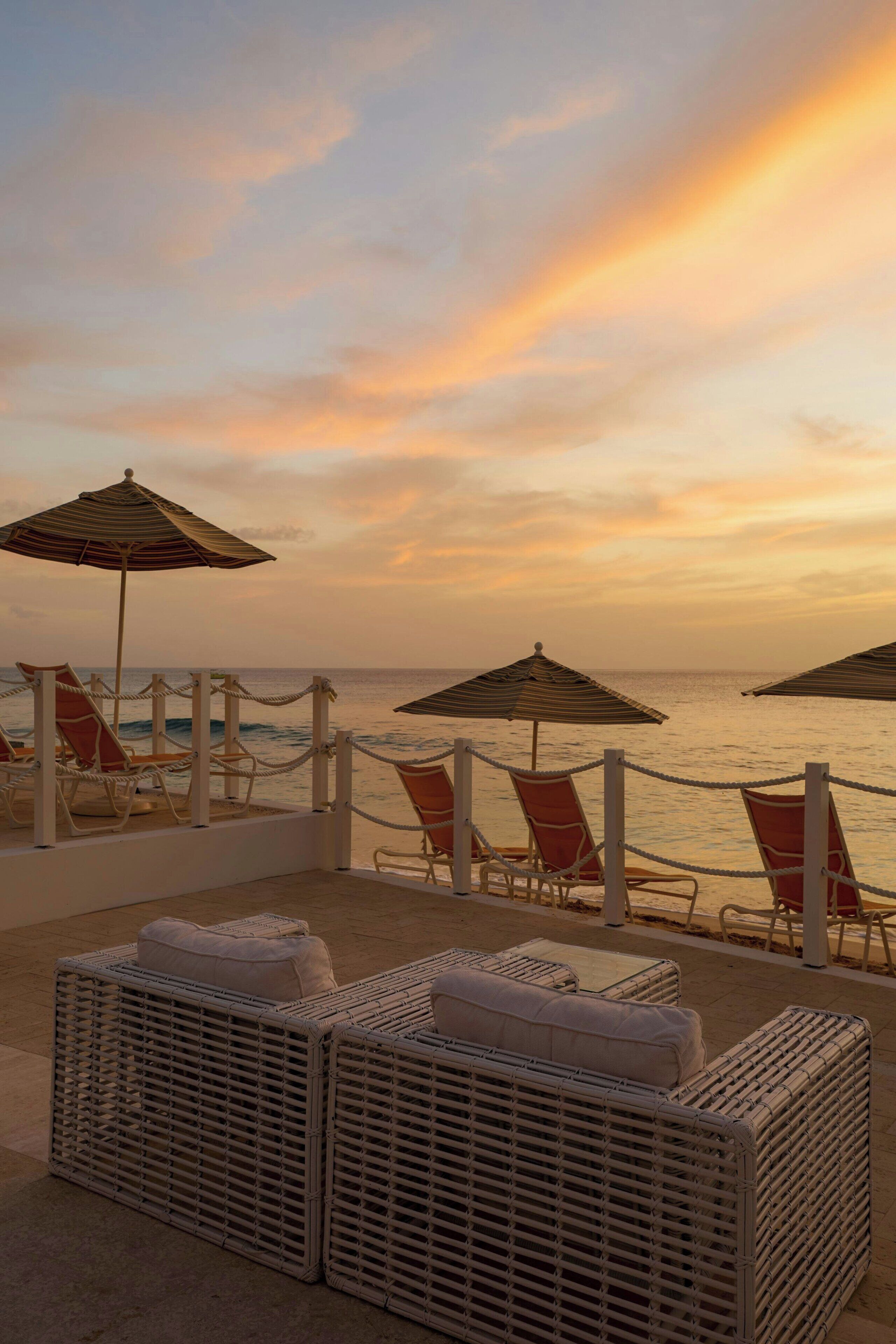A beachside lounge area with wicker furniture and striped umbrellas, overlooking the ocean.