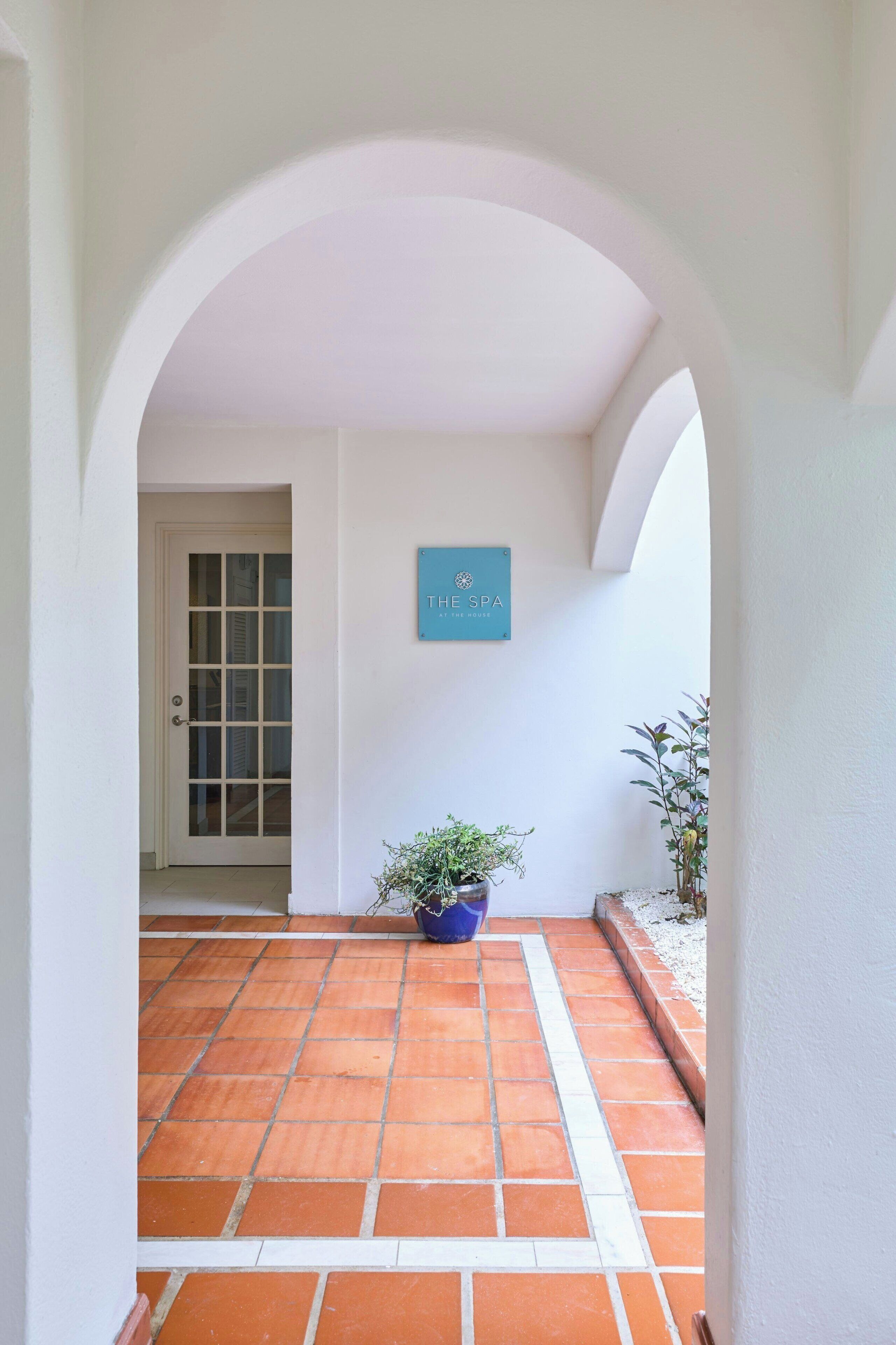A clean, white-walled corridor with a tiled floor leading to a door. A sign above the door reads 'THE SPA AT THE HOUSE.' There are potted plants on either side of the corridor.