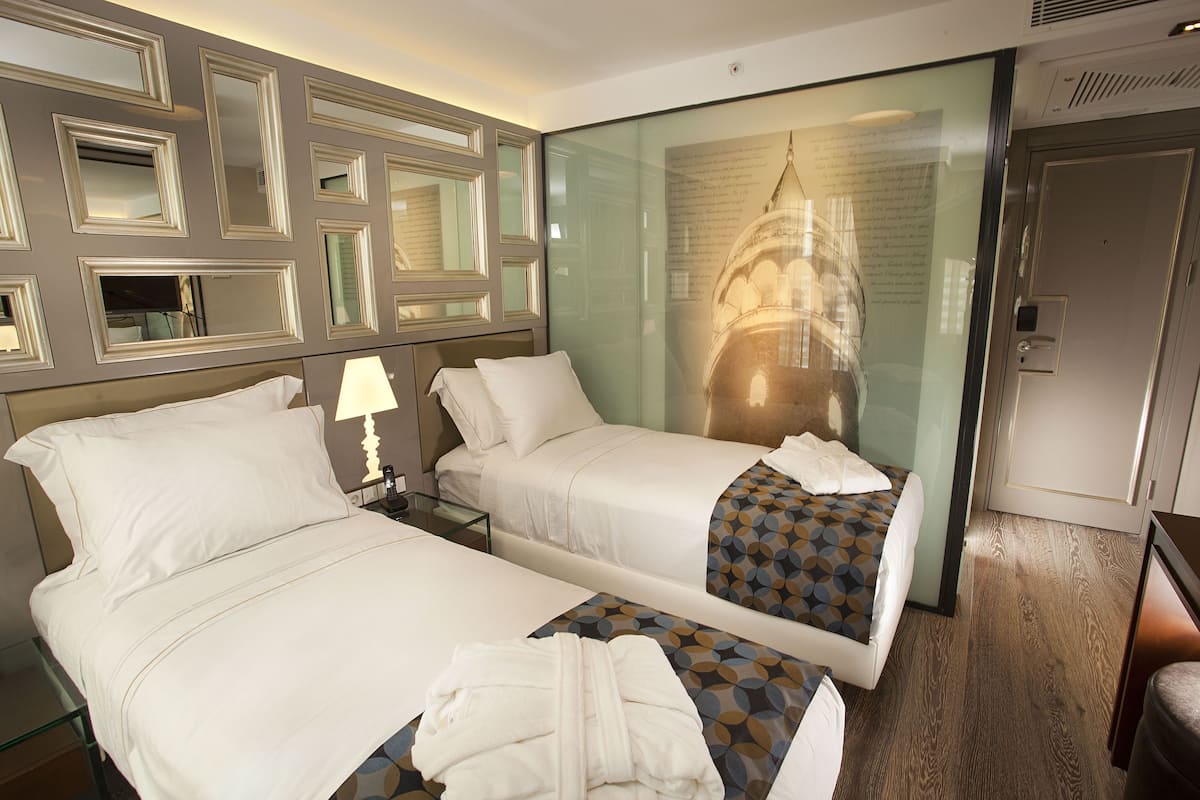 A hotel room with two beds, a glass partition, a lamp, and a view of a building.