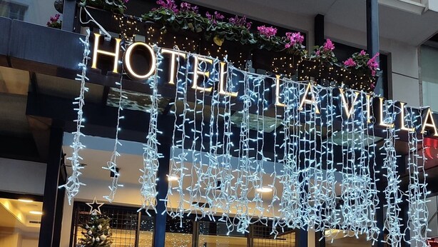 L’entrée d’un hôtel, avec une enseigne lumineuse indiquant « HÔTEL LAVILLA », un sapin de Noël et un paillasson « La Vila ».
