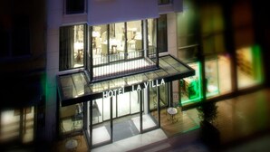 Un hôtel doté d’une façade en verre, d’une salle à manger visible avec des tables et des chaises, et dont l’enseigne « HÔTEL LA VILLA » est clairement affichée.