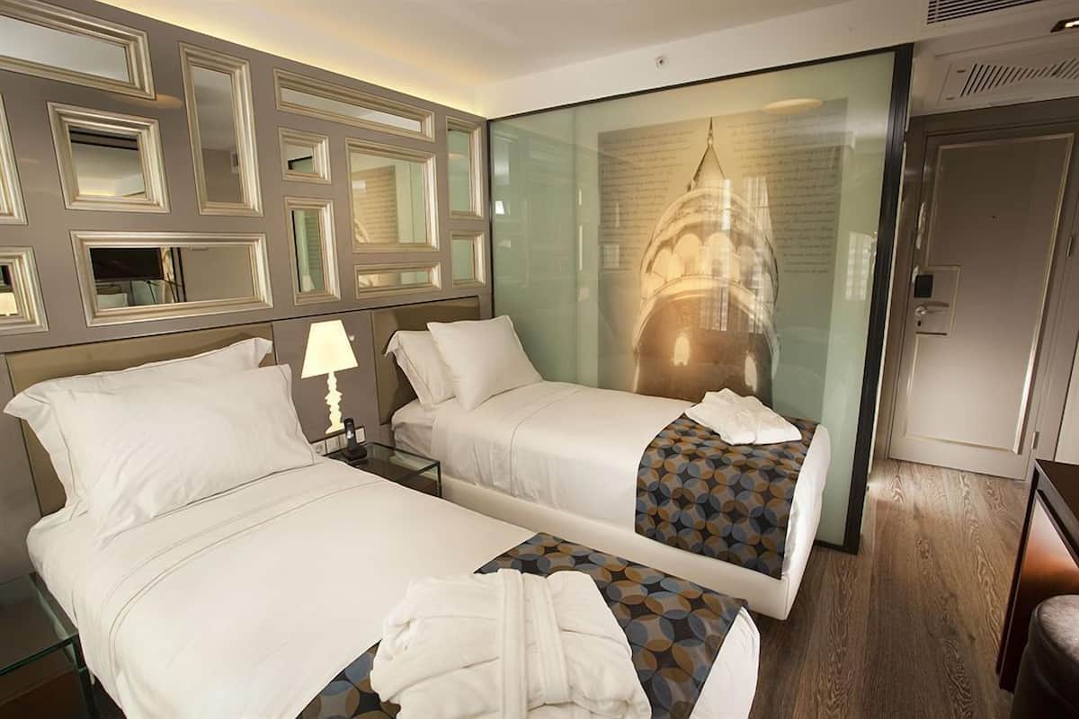 A hotel room with two beds, a glass partition, a lamp, and a view of a building.
