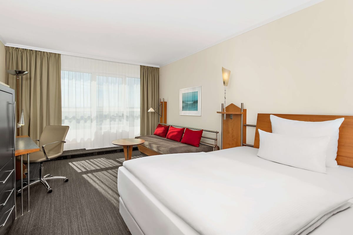 Ein Hotelzimmer mit einem großen Bett, einem Schreibtisch mit Stuhl, einer Couch mit roten Kissen und einem großen Fenster mit Vorhängen.