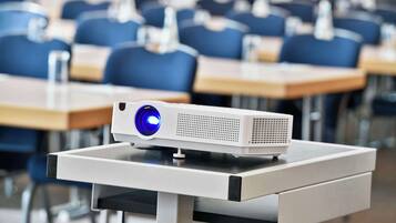 Un aula con filas de sillas azules y un proyector blanco sobre un soporte.