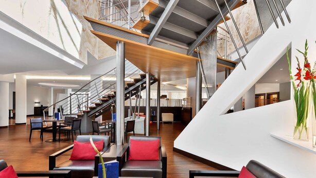 A modern hotel lobby with a wooden floor, black leather sofas with red cushions, a staircase, and a large window. - NH Mannheim-Viernheim (Viernheim)