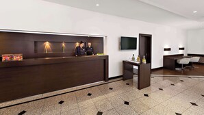 A modern reception area with a dark wood reception desk, two staff members, a flat-screen TV, and a seating area with chairs and a small table. - NH Mannheim-Viernheim (Viernheim)