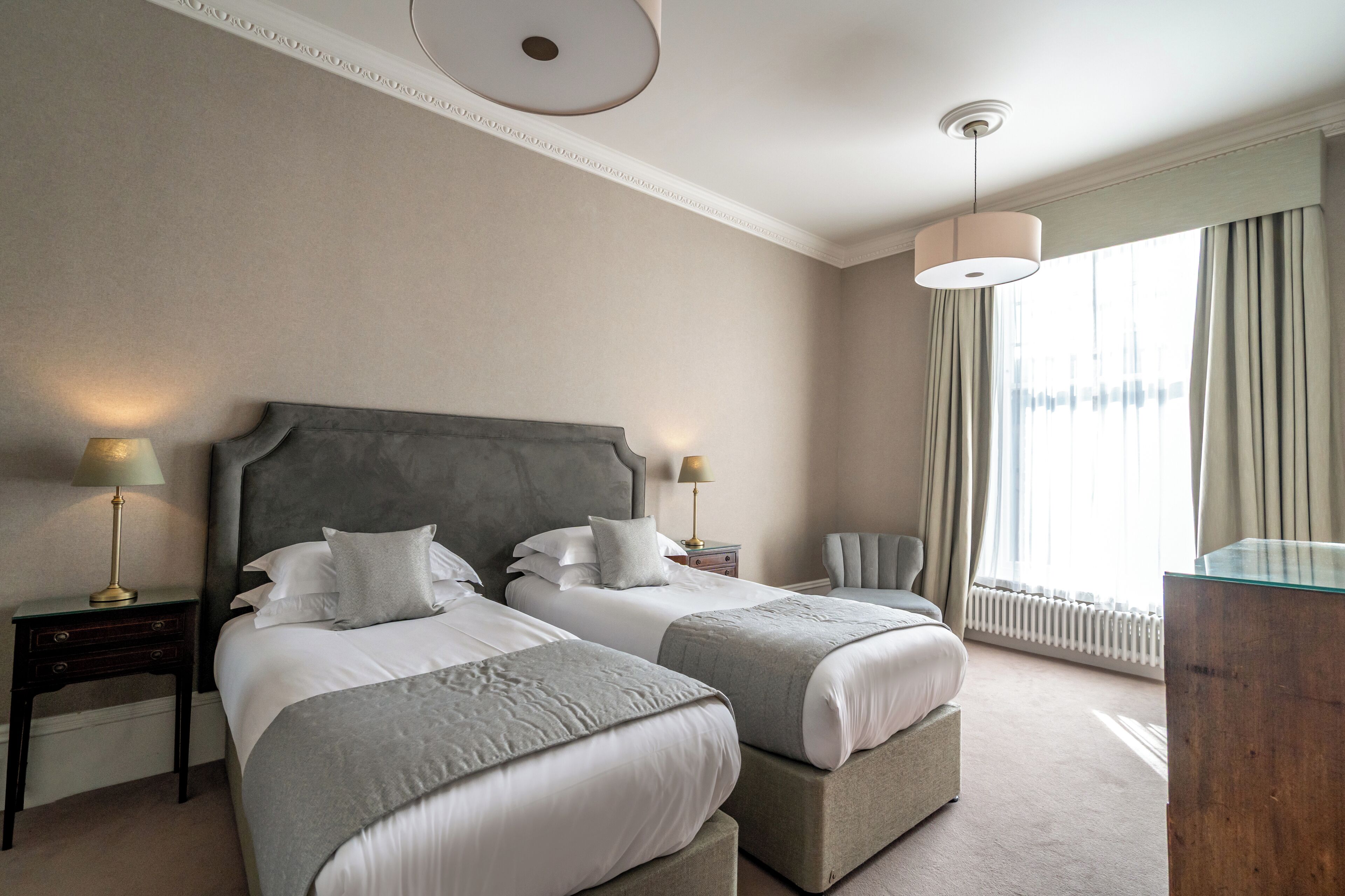Ein Hotelzimmer mit zwei Betten, einem Nachttisch, einem Sessel, einem Schreibtisch und einem großen Fenster mit Vorhängen.