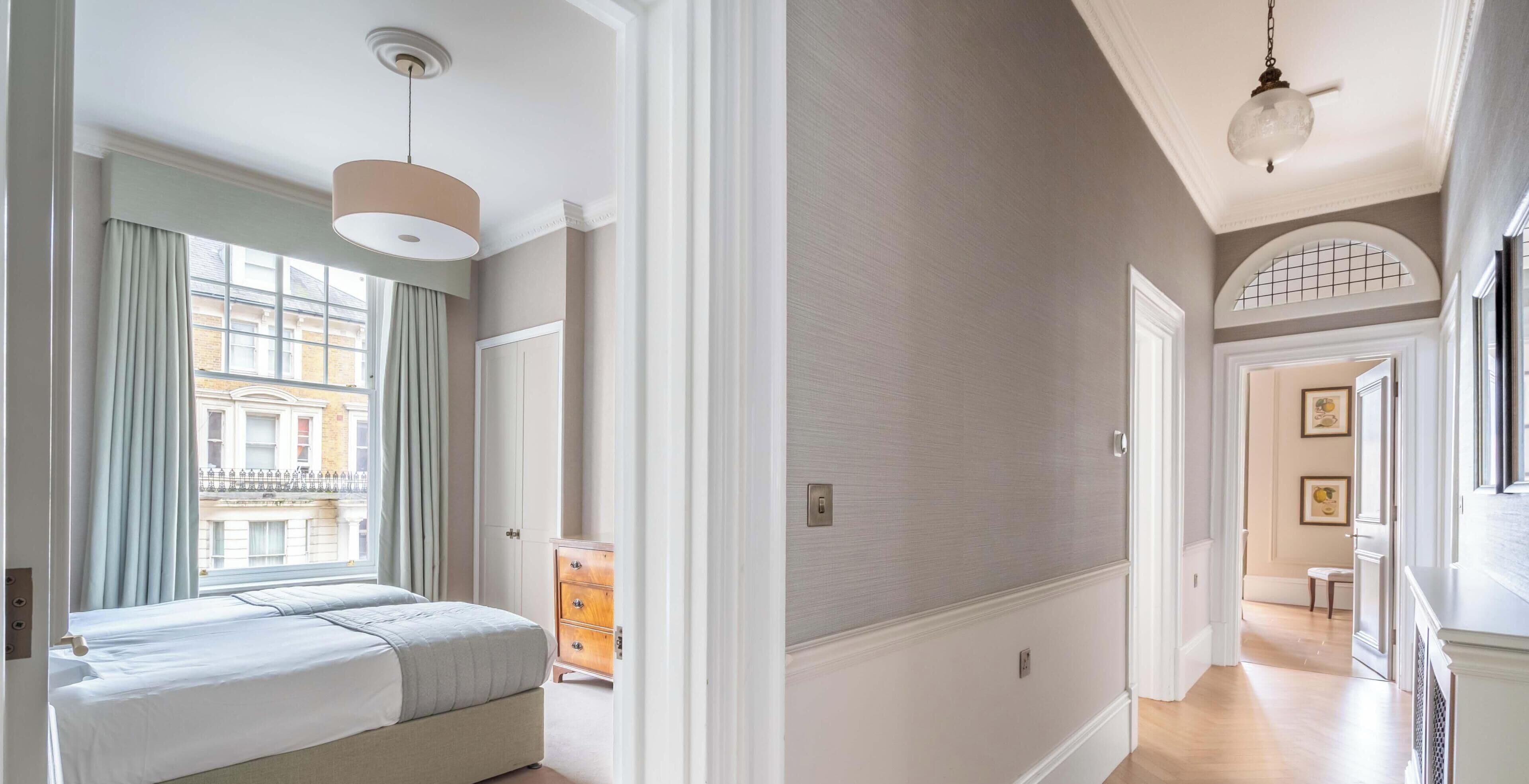 Ein Hotelzimmer mit Bett, Schminktisch und Blick auf die Straße. Das Zimmer hat eine Tür zu einem Flur mit einer Lampe.