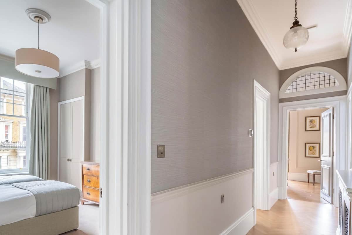 Une chambre d’hôtel avec un lit, une commode et une vue sur la rue. La chambre est dotée d’une porte qui donne sur un couloir éclairé par un luminaire.