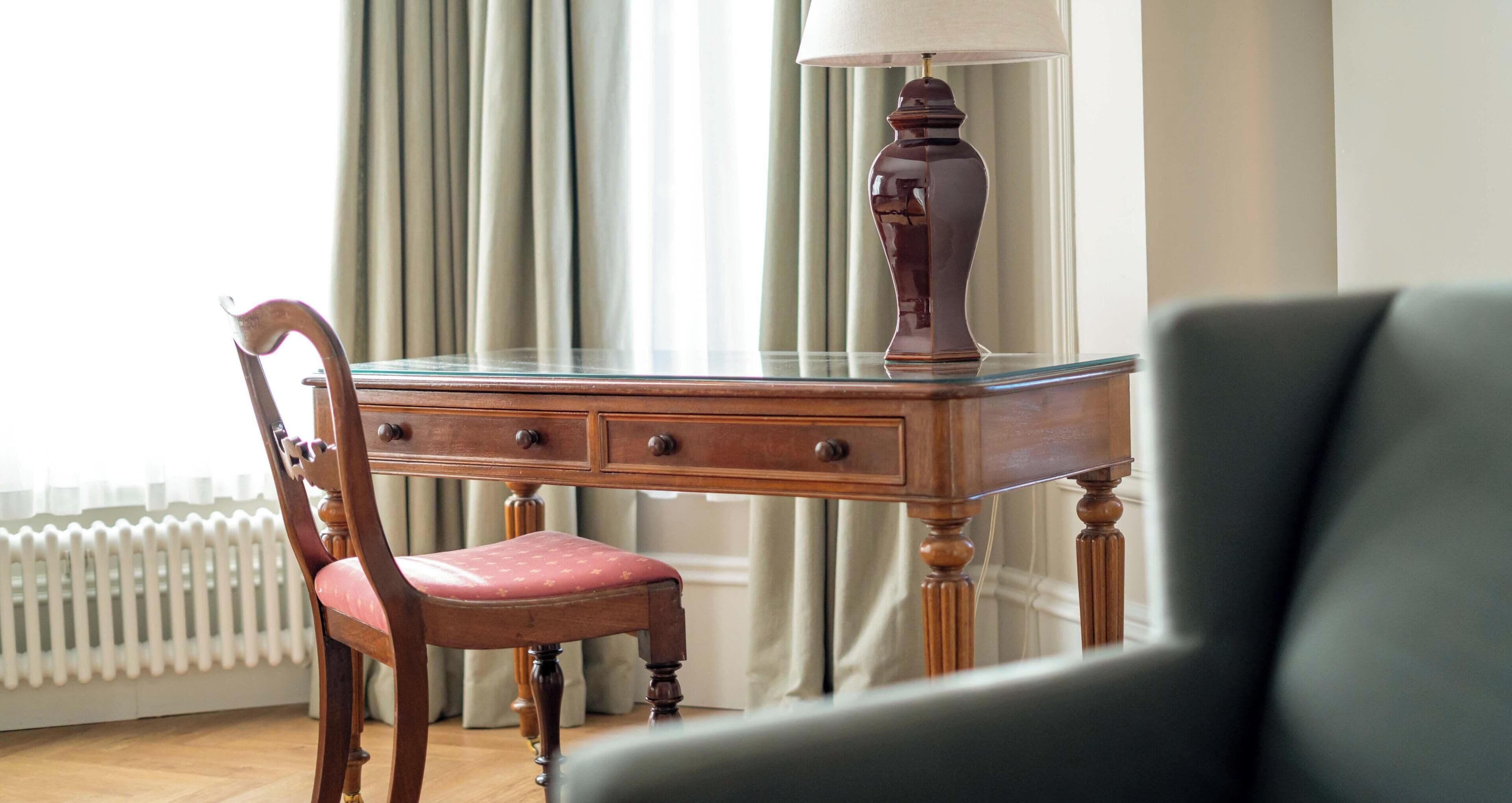 A wooden desk with a glass top, a chair with a cushioned seat, and a lamp.