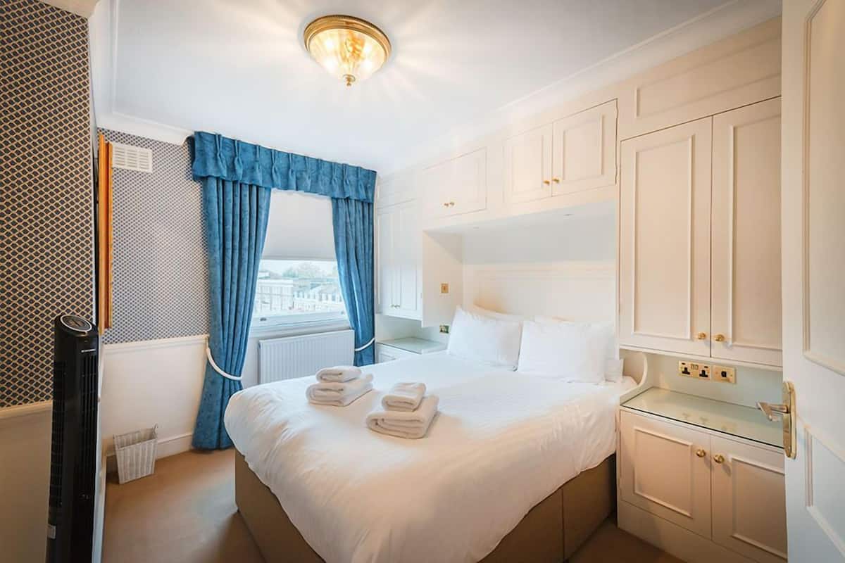 Une chambre d’hôtel avec un grand lit, des murs blancs, une fenêtre avec des rideaux bleus, un radiateur et une petite table de chevet.