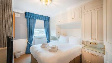 A hotel room with a large bed, white walls, a window with blue curtains, a radiator, and a small bedside table.