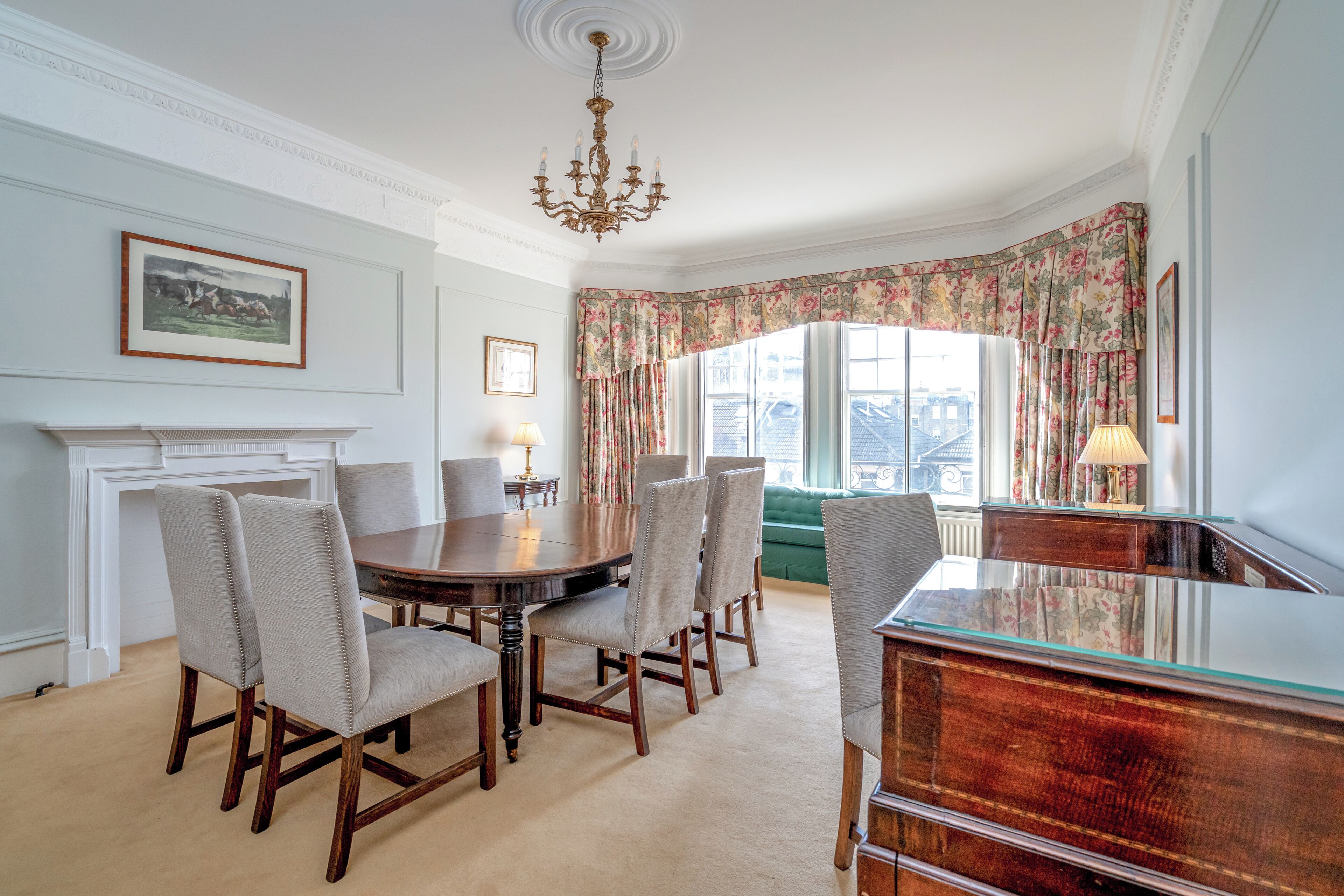 A dining room with a round table, six chairs, a chandelier, a fireplace, and a view of the city through large windows with floral curtains.