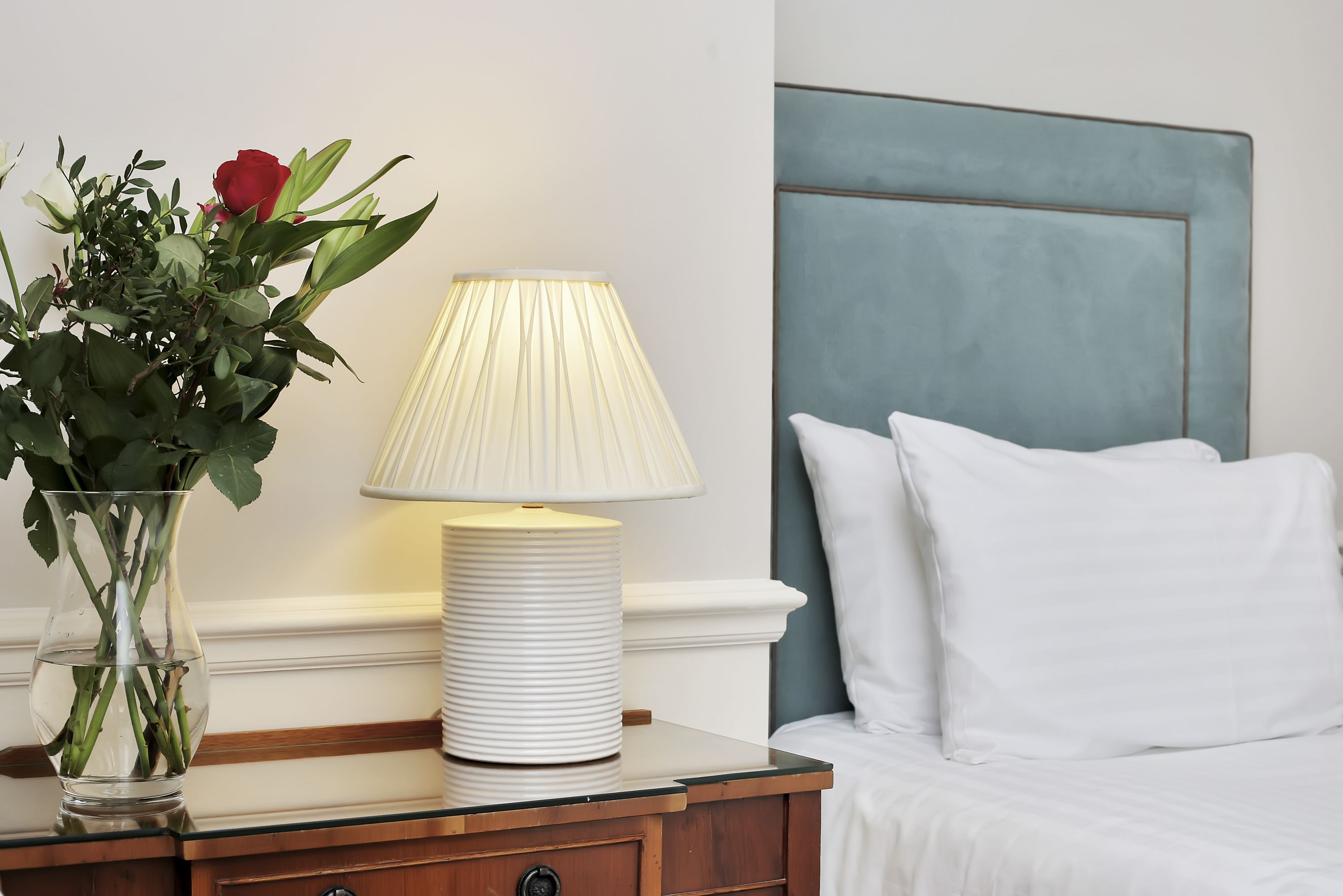 A neatly arranged bedroom with a bedside table, a lamp, a vase with flowers, and a bed with white bedding.