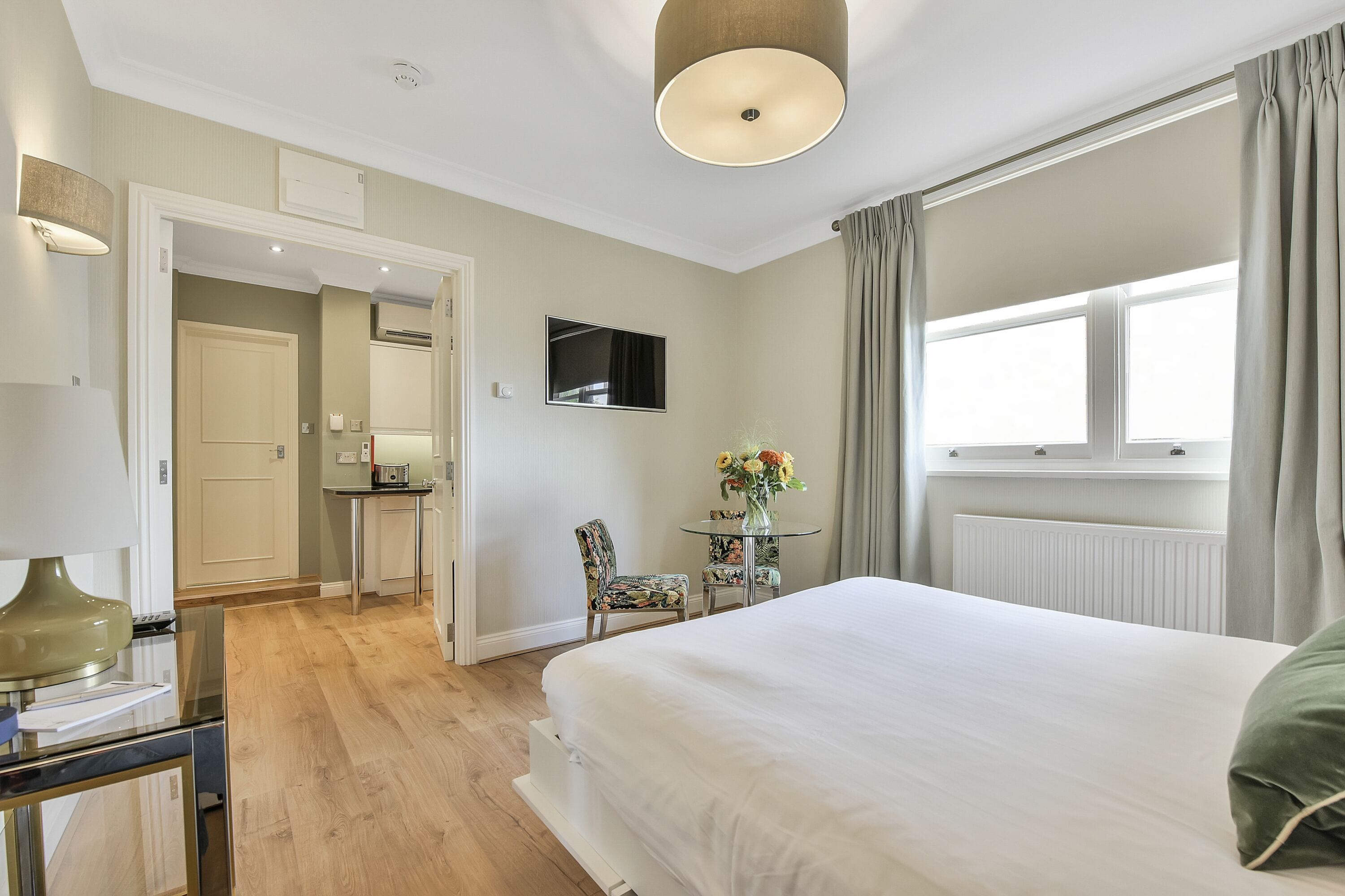 Habitación de hotel con cama, escritorio, silla, una pequeña mesa con flores, televisor y ventana con cortinas.