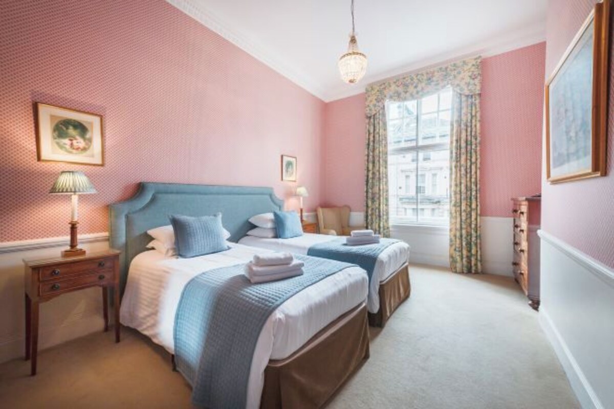 Een hotelkamer met twee bedden, een kroonluchter, een raam met bloemen gordijnen en een ingelijst schilderij aan de muur.