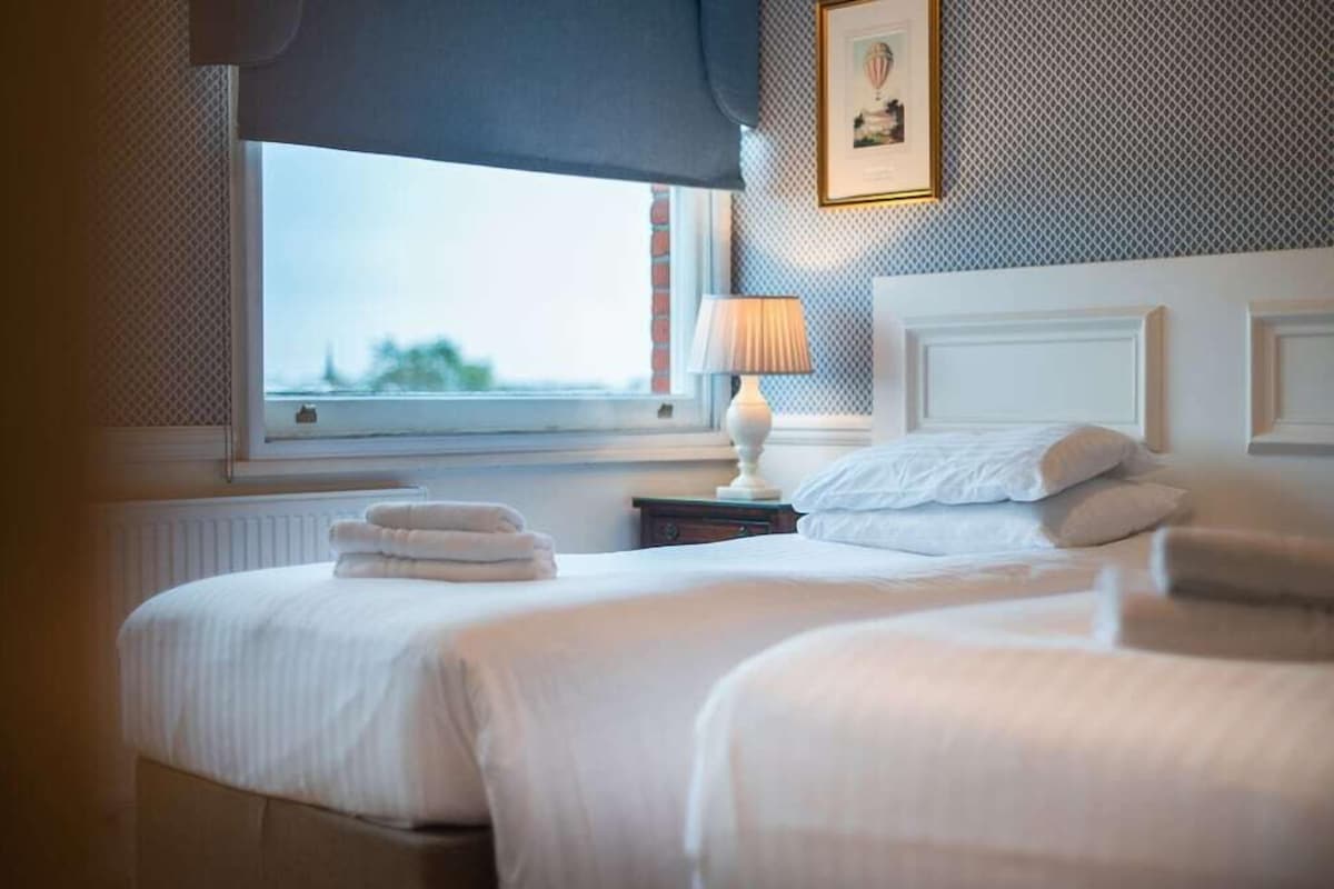 A hotel room with two beds, a window with a view, a lamp on a bedside table, and a framed picture on the wall.