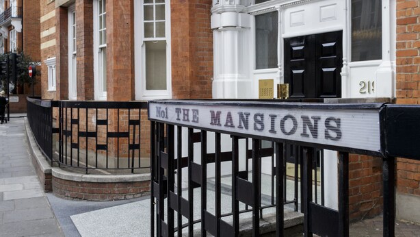 Un immeuble d’angle avec une enseigne indiquant « No 1, The Mansions » et un portail en métal noir.