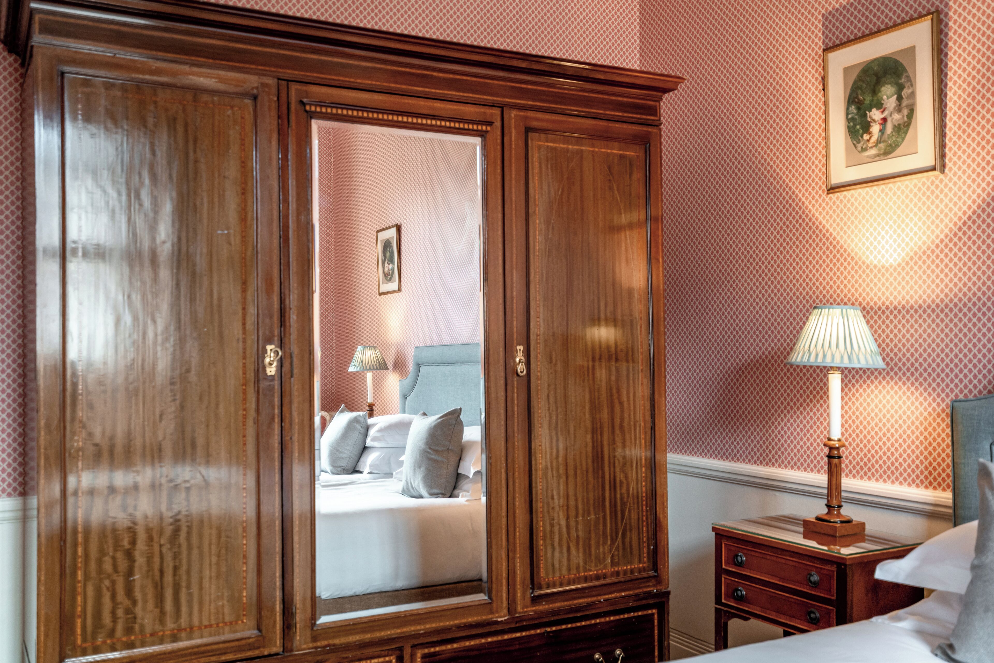 Ein Schlafzimmer mit großem Holzschrank, Nachttisch mit Lampe und einem gerahmten Bild an der Wand.