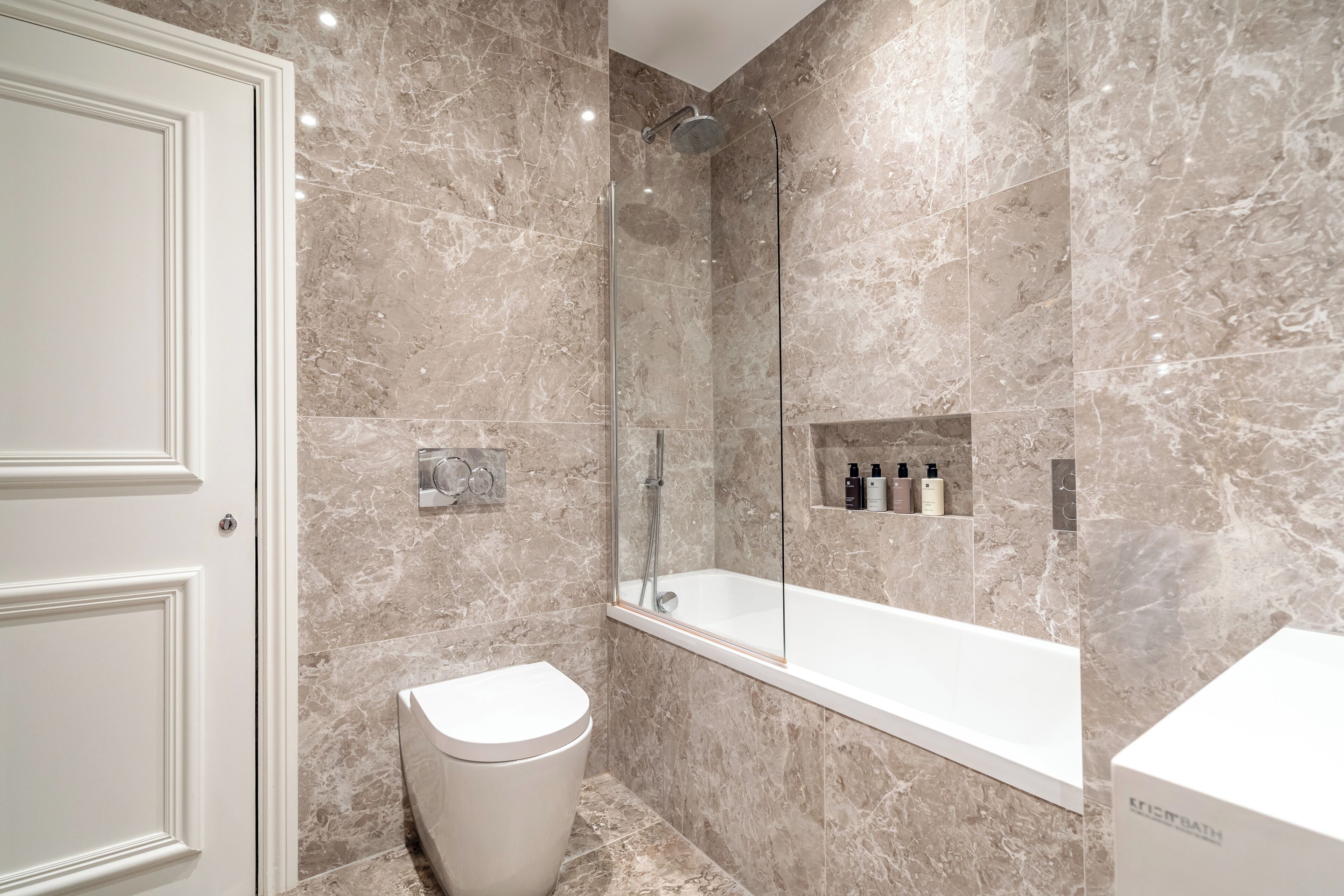 A modern bathroom with a marble wall, a glass-enclosed shower, a white toilet, and a white bathtub.