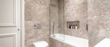 A modern bathroom with a marble wall, a glass-enclosed shower, a white toilet, and a white bathtub.