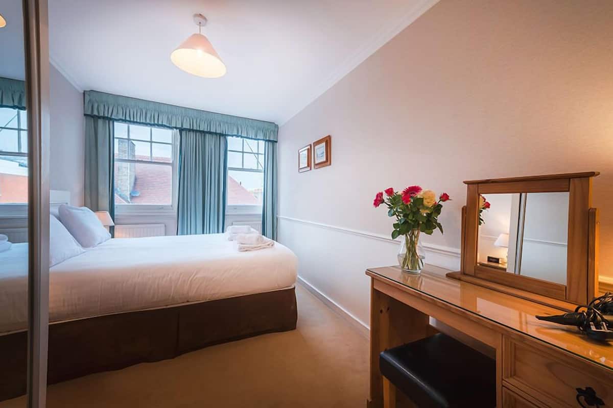 Une chambre d’hôtel avec un lit, une table de chevet avec un miroir, un vase de fleurs et une fenêtre avec des rideaux.