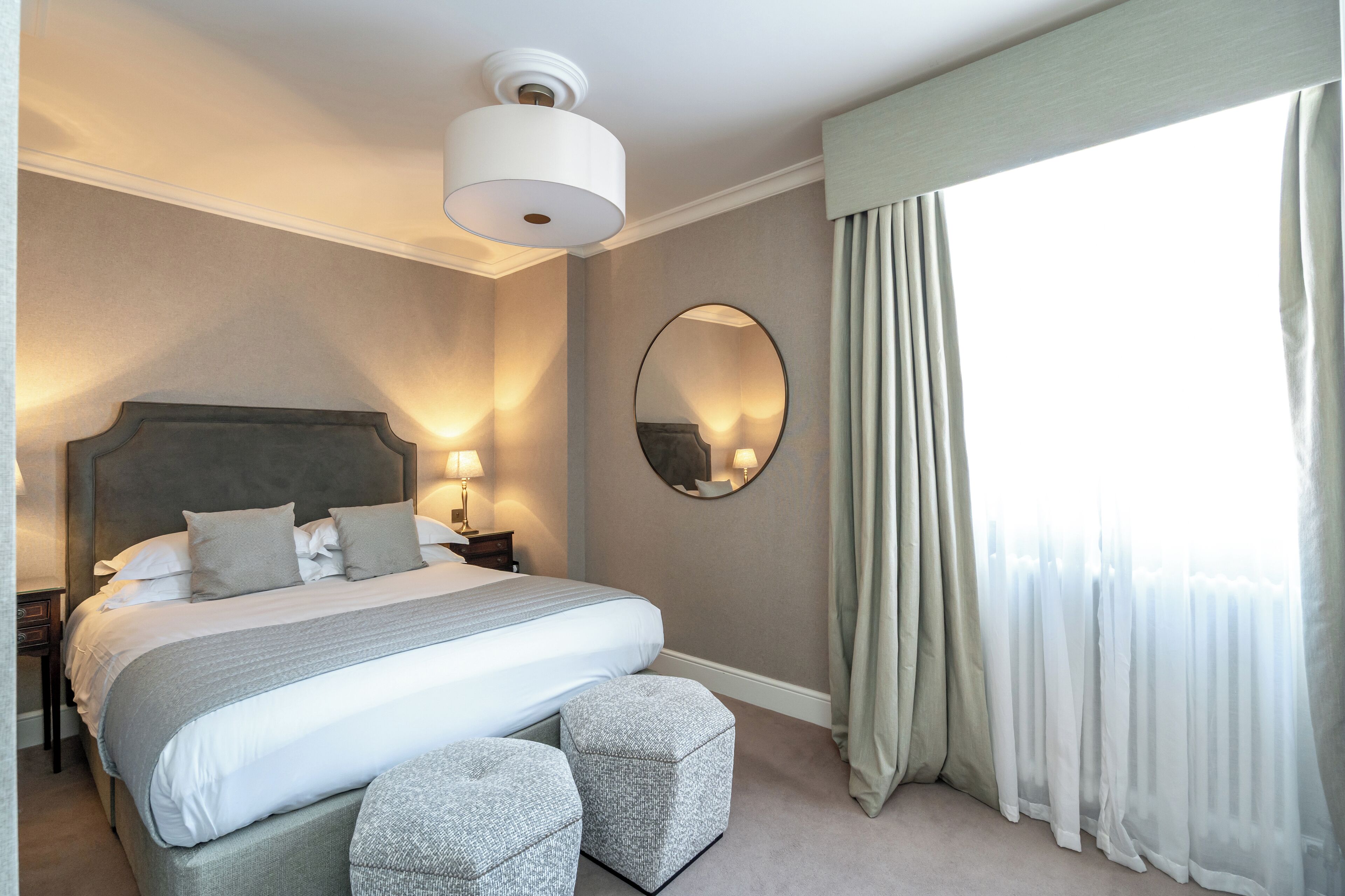 A neatly arranged bedroom with a large bed, bedside lamps, a round mirror, and a window with sheer curtains.