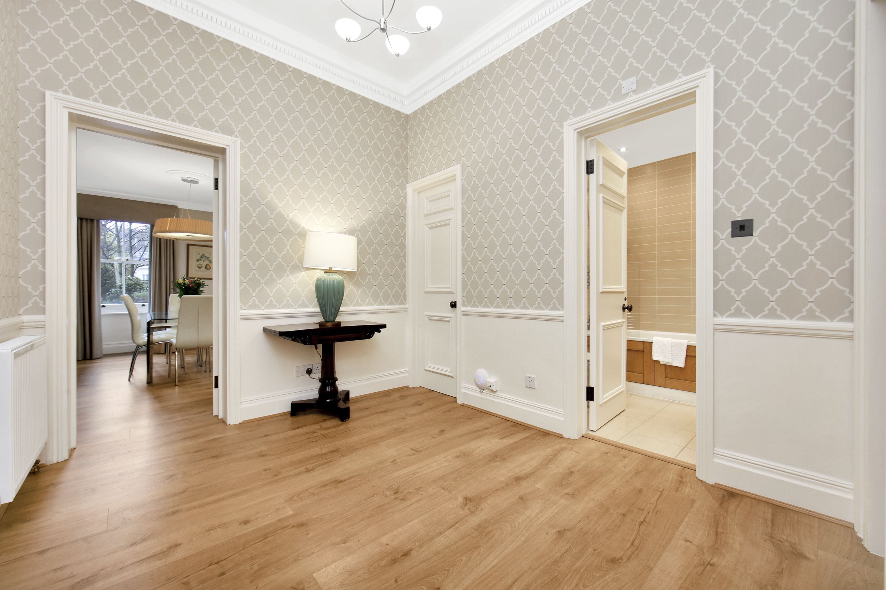 A hallway with wooden flooring, white wainscoting, and patterned wallpaper. There is a table with a lamp and chairs, and a door leading to a bathroom.