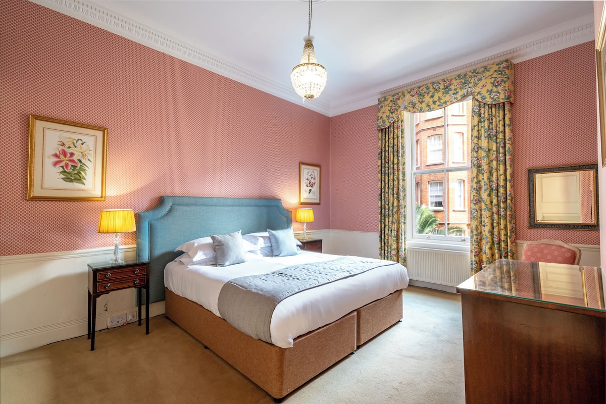 Une chambre à coucher avec un grand lit, deux tables de chevet, une commode, un lustre et une fenêtre ornée de rideaux à motifs floraux.