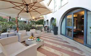 Un espace extérieur aménagé en terrasse, avec des meubles en osier, une table sur laquelle sont disposés des fruits et un verre, et un grand parasol.