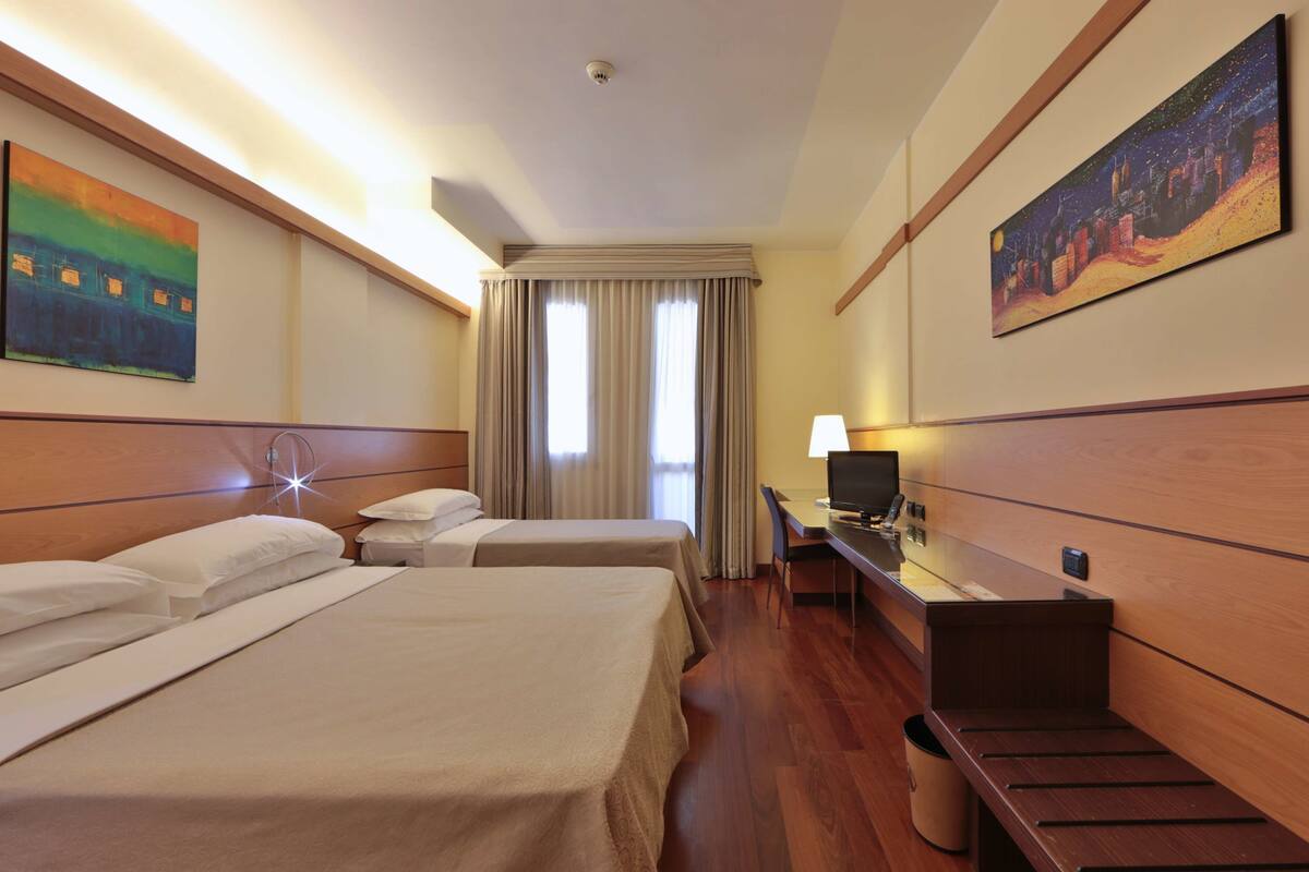 Camera d'albergo con due letti, una scrivania con un computer, una televisione e opere d'arte alle pareti.
