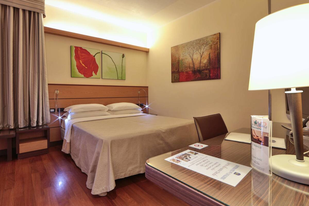 Una camera d'hotel con un letto, una scrivania con una lampada e opere d'arte alle pareti.