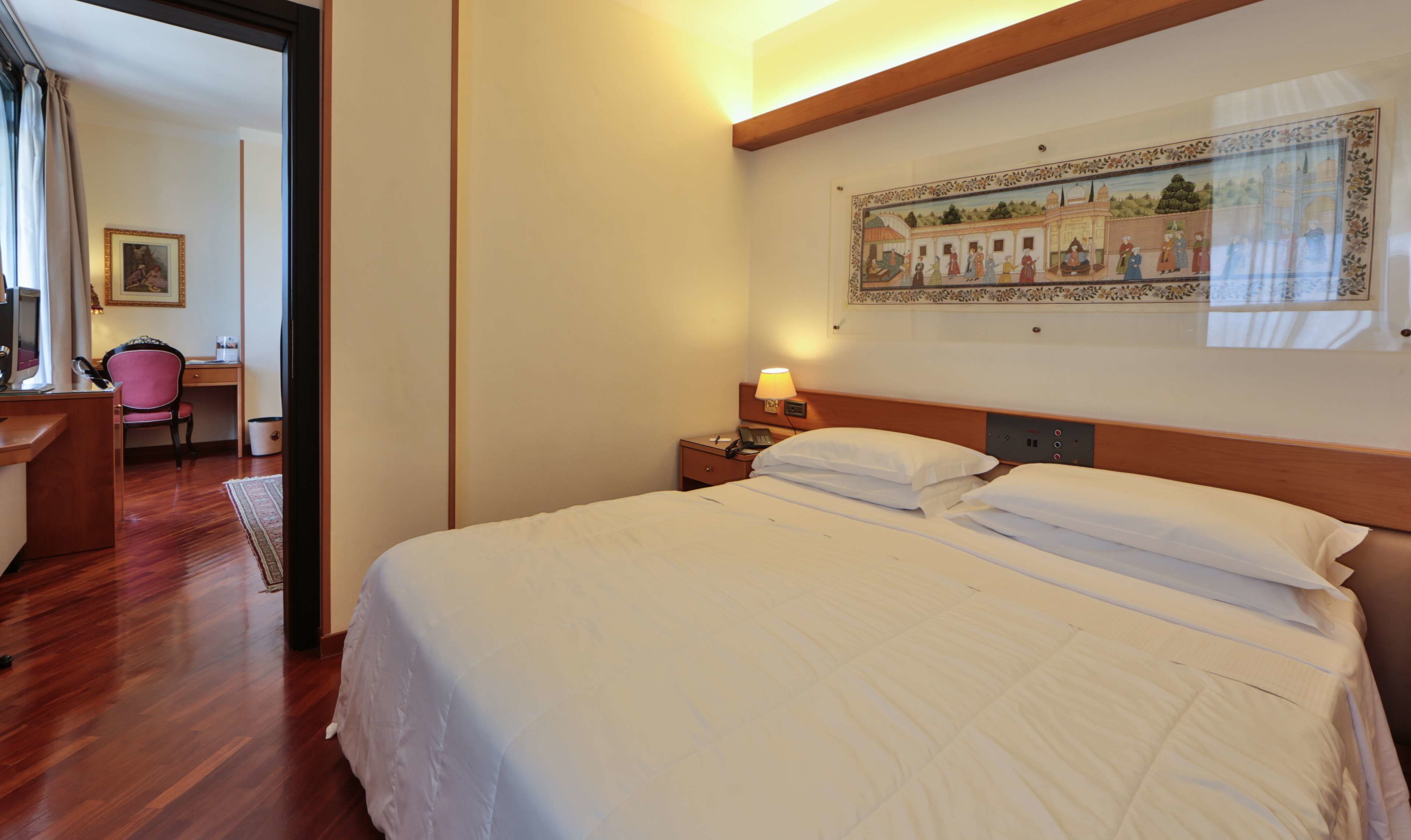 A hotel room with a large bed, a desk with a chair, a lamp, and a painting on the wall.