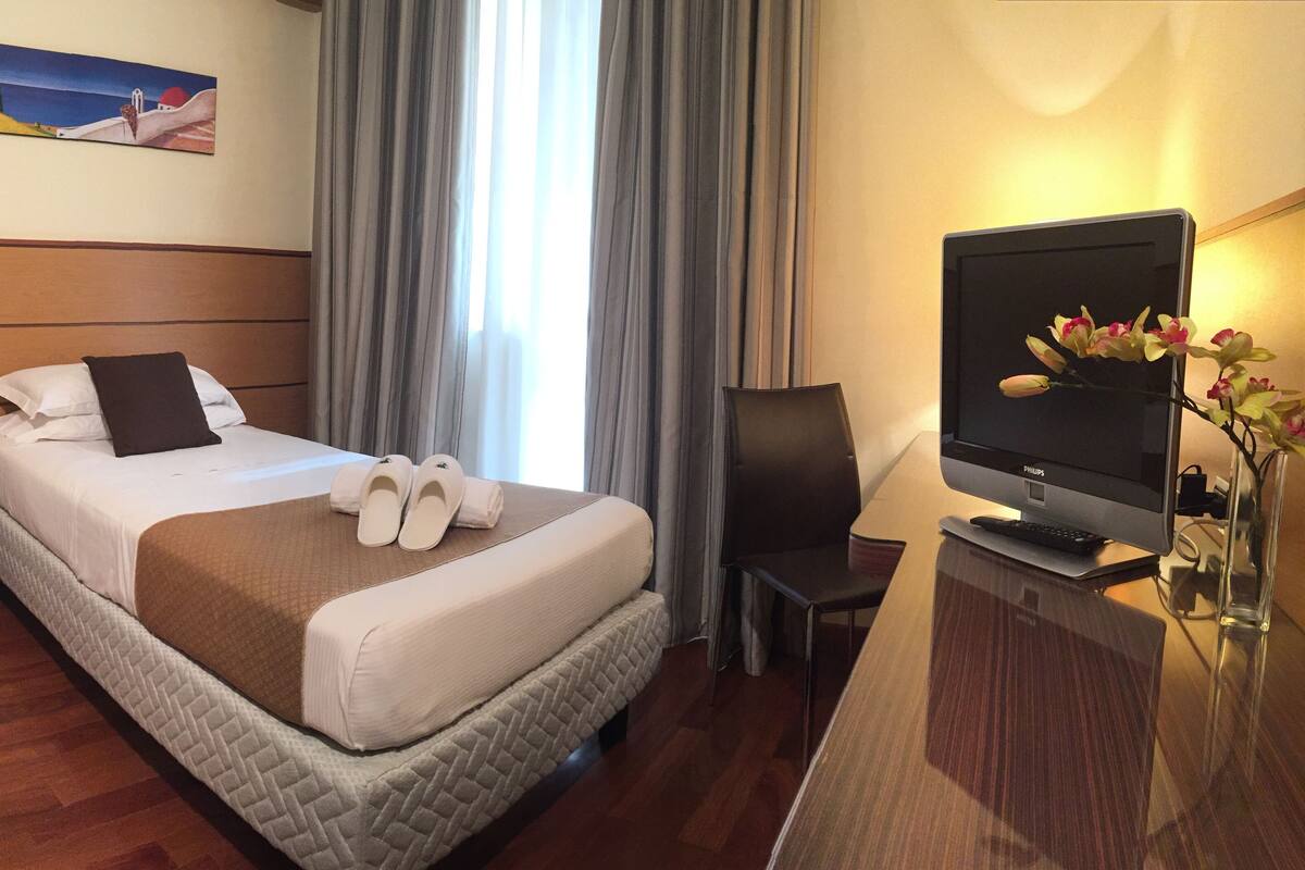Una camera d'albergo con un letto, una TV, una scrivania con un computer e un quadro appeso al muro.