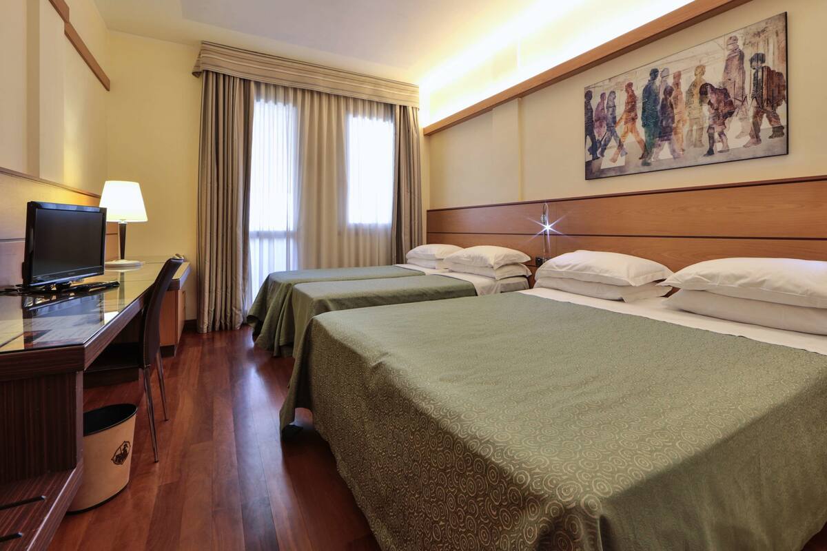 Una camera d'albergo con due letti singoli, una scrivania con televisore, una lampada e un quadro appeso al muro.