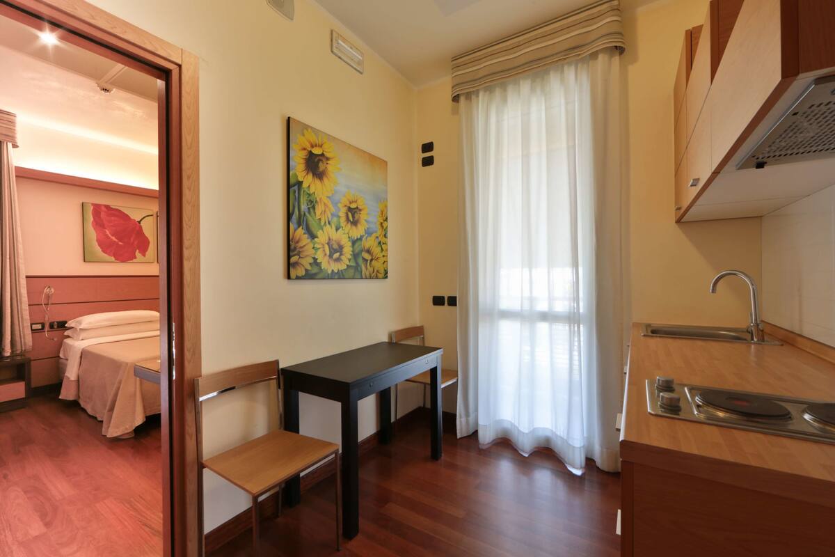 Camera d'albergo compatta con un letto, un tavolino, una sedia, un quadro, una finestra con tende e angolo cottura.
