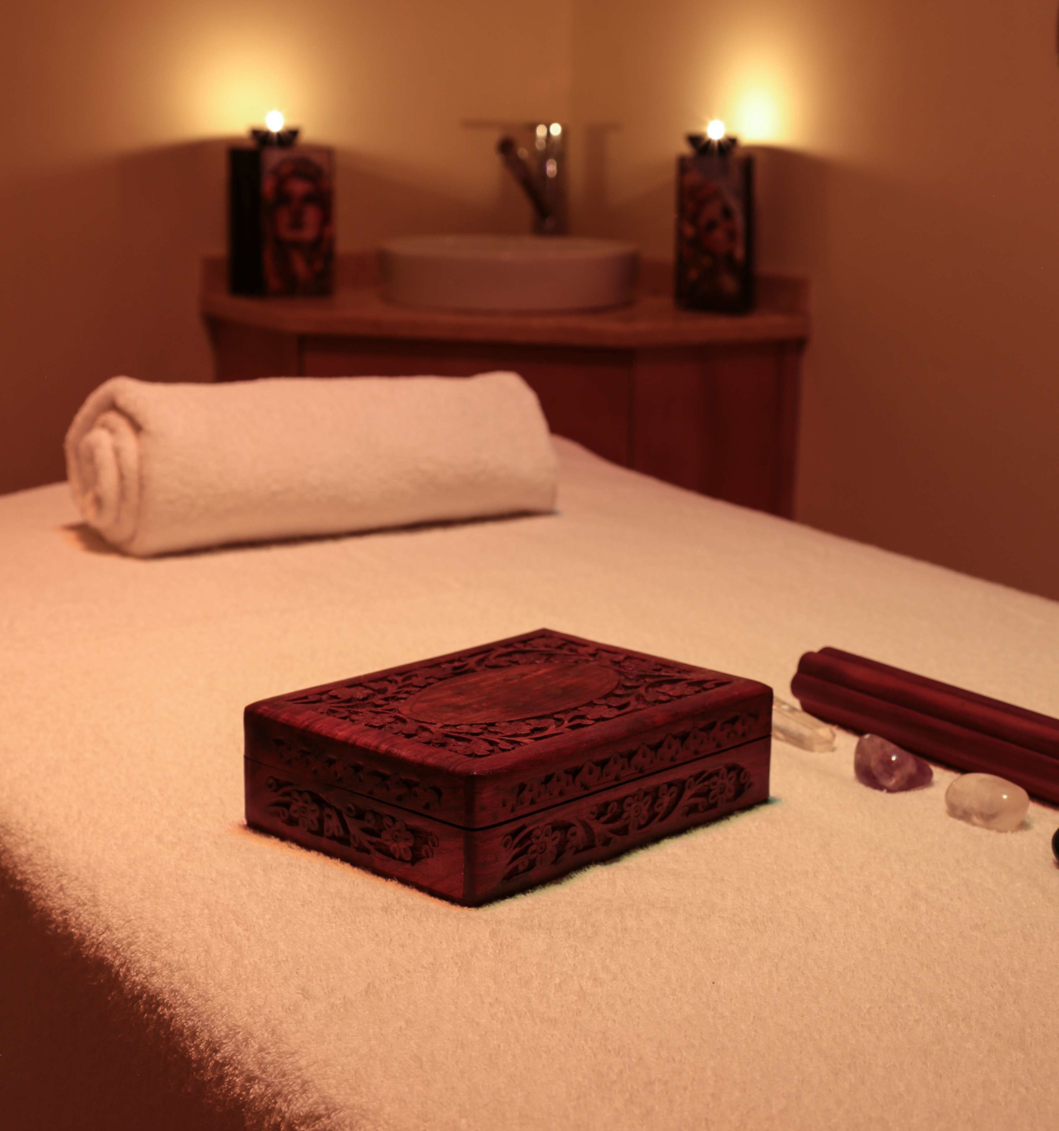 A neatly made bed with a rolled white towel, a wooden box with intricate carvings, and a few stones on the bed.